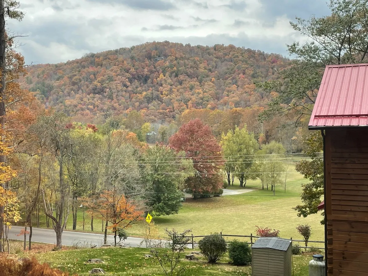 A sweeping view of vibrant autumn foliage, rolling hills, an expansive green field, and a distant mountain range. The open landscape invites outdoor activities such as hiking, picnicking, and play, with a serene, family-friendly atmosphere.