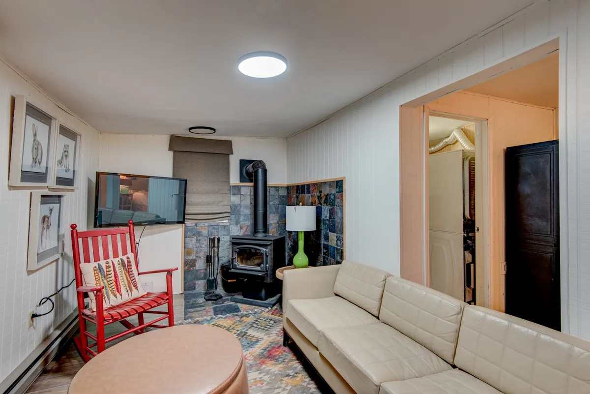 A welcoming lower den features a classic wood stove, a colorful rug, a modern sofa, and a red rocking chair arranged for comfort and socializing. The space exudes warmth and relaxation, making it ideal for family gatherings, movie nights, or quiet moments by the fire.