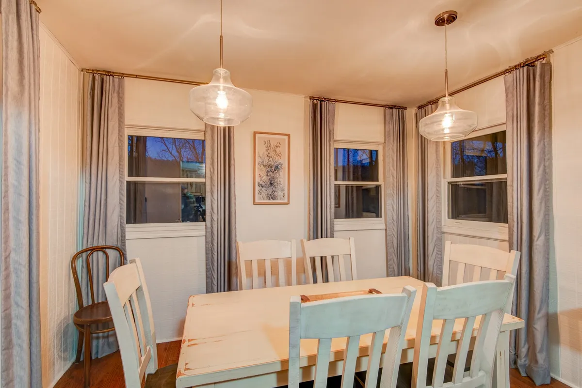 This image features a dining area with an elegant table, hanging light fixtures, window curtains, and framed artwork. The table is set to accommodate a group, emphasizing communal meals and togetherness in a warm, softly lit atmosphere.