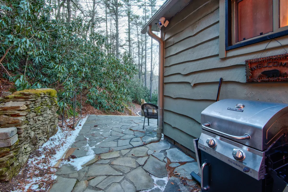 A stone patio with a grill, chairs, lush surrounding greenery, and a welcoming sign, offering a cozy spot to relax after outdoor adventures. The space feels inviting and is connected to the outdoors.