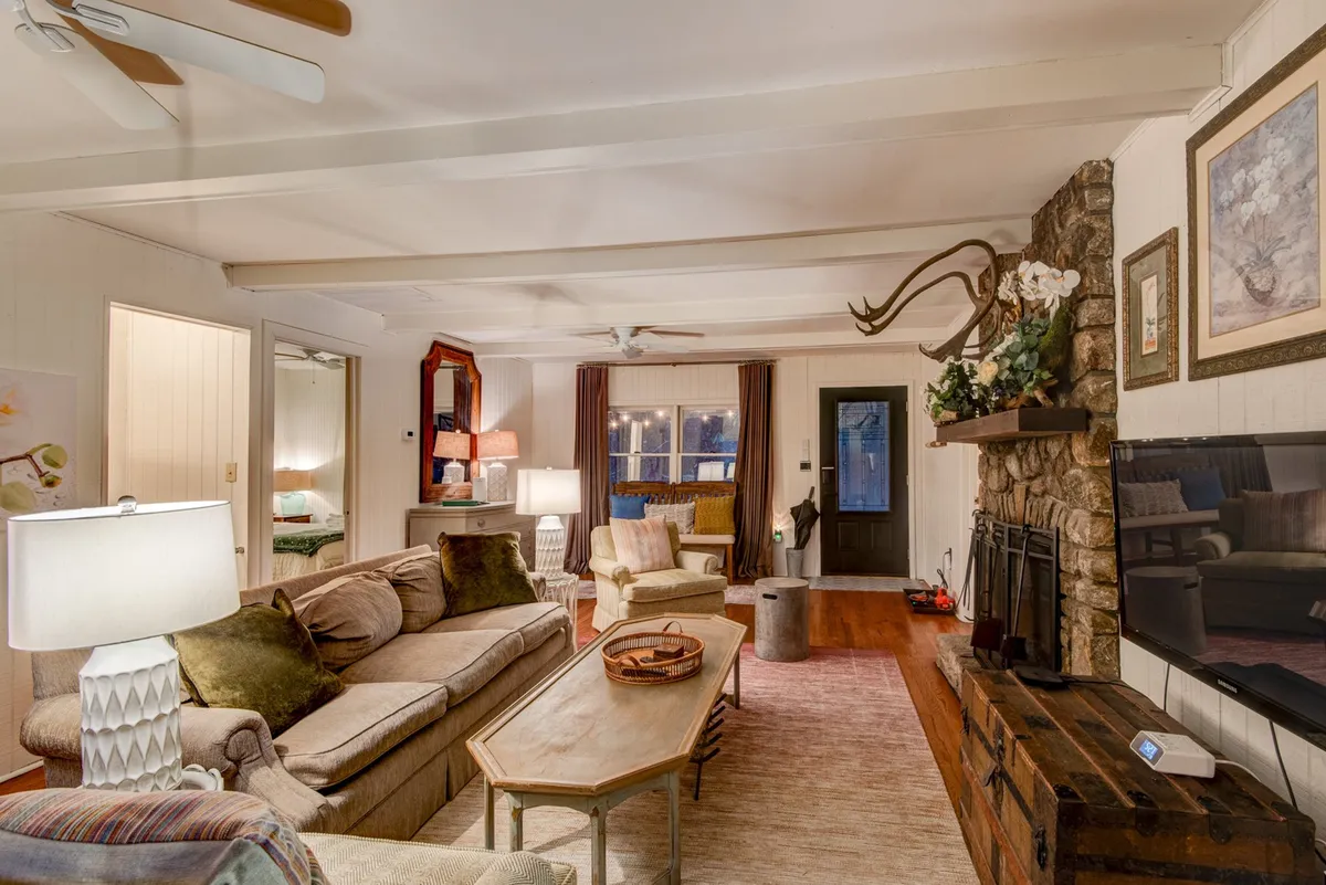 The image captures a spacious living area with a stone fireplace as the focal point, flanked by a comfortable sofa, antler décor, and large windows with curtains that let in natural light. The arrangement of seating encourages socializing and relaxation, and the warmth of the fire makes the space feel inviting.