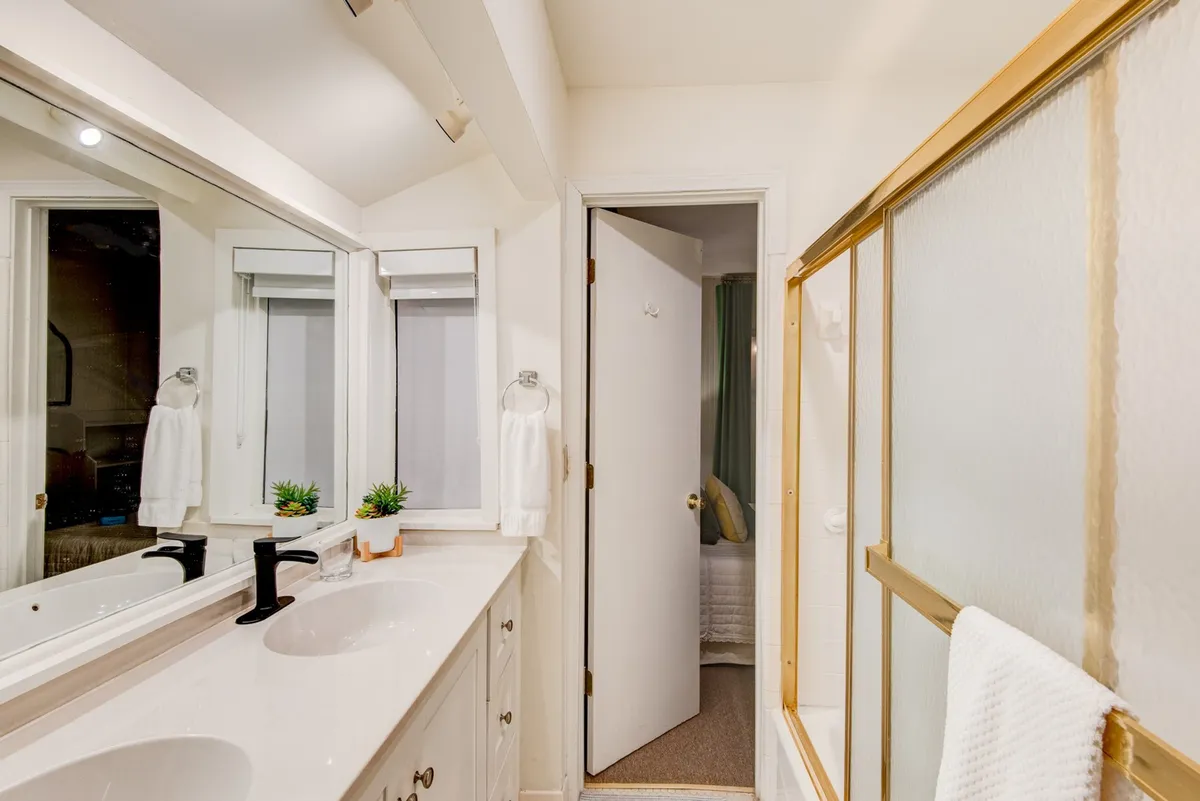 A bright, inviting jack-and-jill bathroom with a double sink vanity, large mirror, glass shower enclosure, fluffy towels, and a touch of greenery. The space feels organized, spacious, and ideal for shared routines.