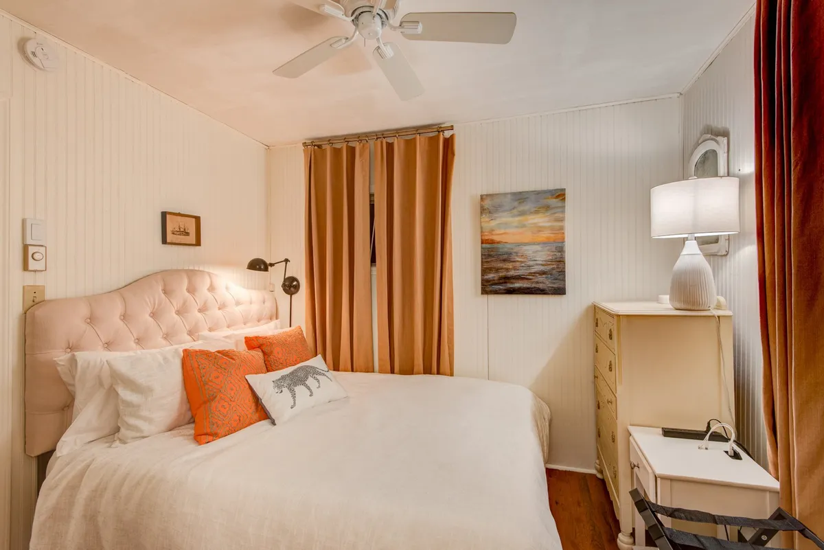A serene bedroom featuring a bed with a pink headboard, orange accent pillows, a nightstand with a lamp, soft curtains, and a calming painting of a seascape. The inviting atmosphere is enhanced by natural light filtering through the curtains, creating a perfect space for relaxation and restful sleep.